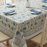 A close-up, high-angle view of a dining table featuring a stylised floral block-print tablecloth in a blue, teal and warm sand. The table is set with white plates and slate-blue napkins.