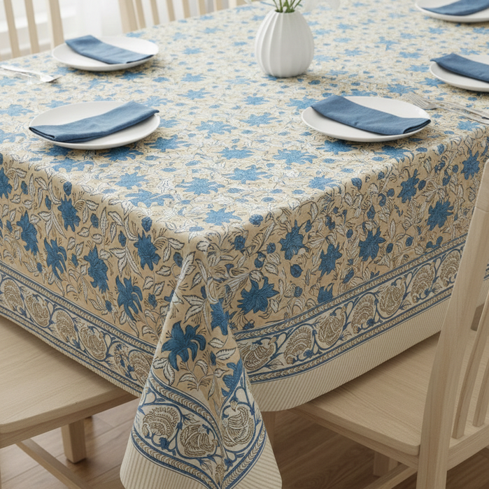 A close-up, high-angle view of a dining table featuring a stylised floral block-print tablecloth in a blue, teal and warm sand. The table is set with white plates and slate-blue napkins.