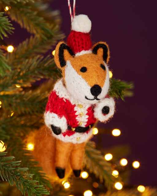 A felt fox ornament wearing a Santa hat and a knitted sweater hangs on a Christmas tree.