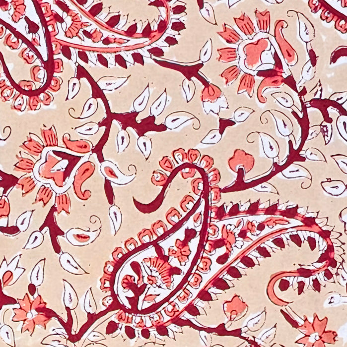 A close-up of a printed cotton tablecloth featuring red and orange paisley and floral motifs on a warm peach background.
