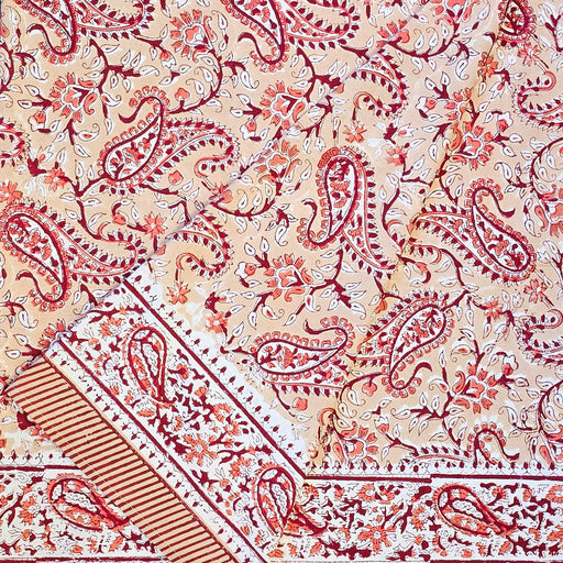 A folded section of a printed cotton tablecloth featuring red and orange paisley and floral motifs on a warm peach background, with a decorative border running horizontally across the lower portion.