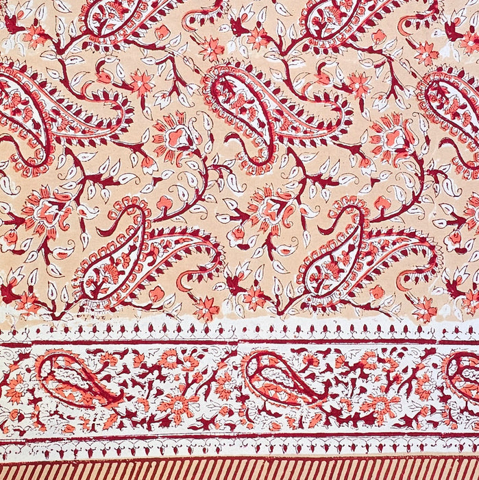 A close-up of a printed cotton tablecloth featuring red and orange paisley and floral motifs on a warm peach background, with a decorative border running horizontally across the lower portion.