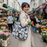 A young woman smiling in London's Columbia Road Flower Market, carrying a large slate grey quilted cotton tote bag with a white crane print. The bag is filled with fresh eucalyptus and tulips, showing its spacious 15cm gusset and comfortable over-the-shoulder fit