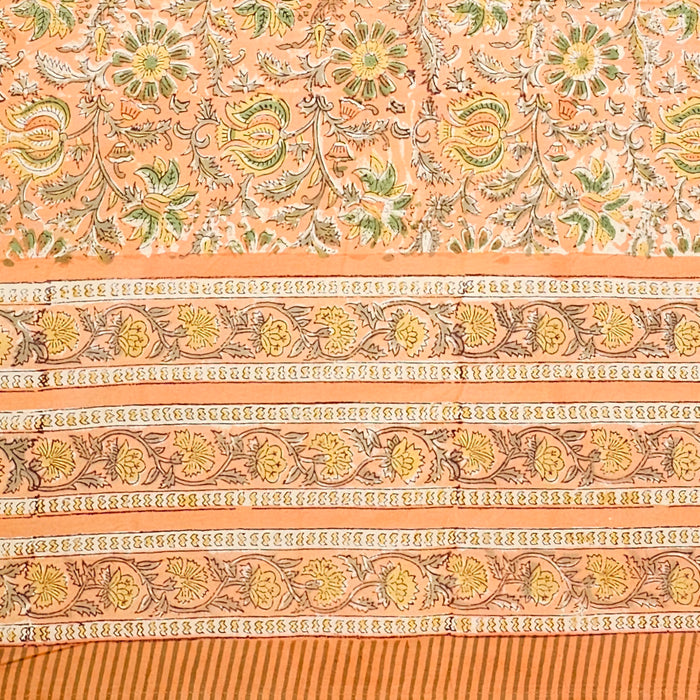 A close-up of a peach-colored fabric tablecloth with an all-over floral block print in green, yellow, and brown.  There is a triple layer border and the fabric is edged with coordinating striped ticking.