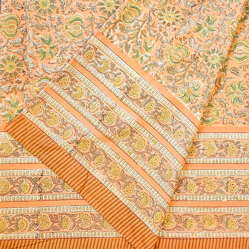 A close-up of a peach-colored fabric tablecloth with an all-over floral block print in green, yellow, and brown, featuring a wide decorative border.  The fabric is folded.
