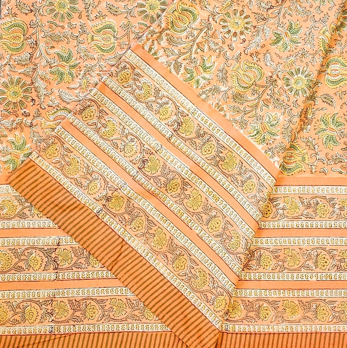 A close-up of a peach-colored fabric tablecloth with an all-over floral block print in green, yellow, and brown, featuring a wide decorative border.  The fabric is folded.