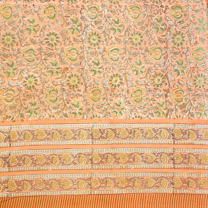 A close-up of a peach-colored fabric tablecloth with an all-over floral block print in green, yellow, and brown, featuring a wide decorative border.