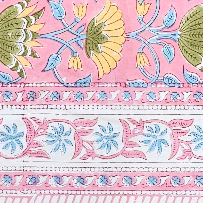 A close-up of a pink patterned tablecloth featuring hand-painted style floral motifs in yellow, green, and light blue, with decorative horizontal borders.