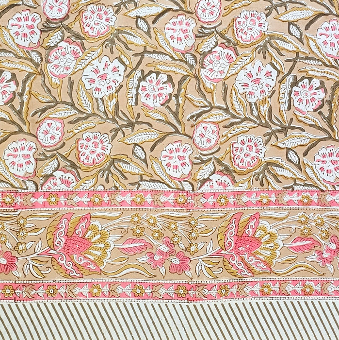 A section of a hand-block-printed tablecloth features a repeating floral and leaf motif in pink, white, and brown against a warm camel background, with a decorative border along the bottom.