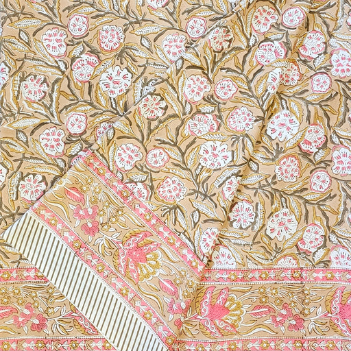 A folded section of a hand-block-printed tablecloth features a repeating floral and leaf motif in pink, white, and brown against a warm camel background, with a decorative border along the bottom.
