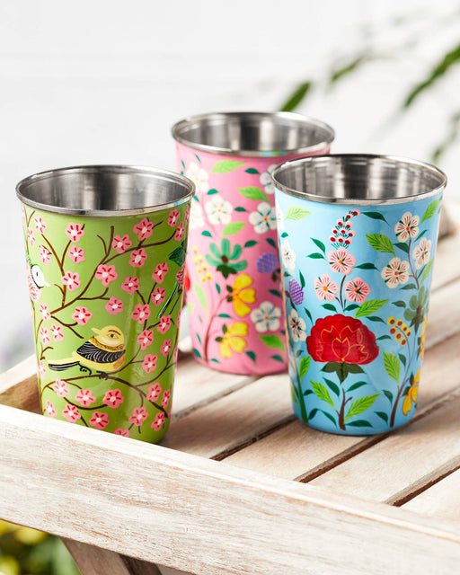 Three colourful hand-painted stainless steel and enamel tumblers with floral designs on a wooden surface