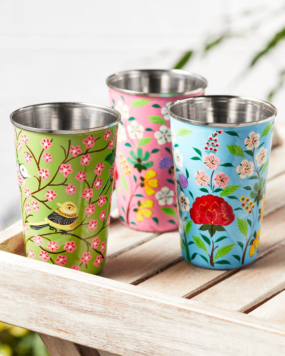 Three colourful hand-painted stainless steel and enamel tumblers with floral designs on a wooden surface