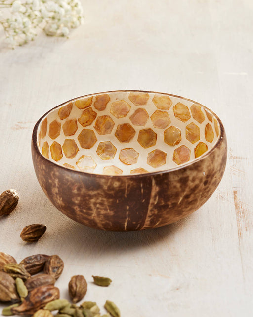 Coconut shell bowl with honeycomb pattern on a light wooden surface with cardamom pods.