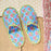 Pair of floral-patterned slippers on a wooden floor with a textured white rug in the background.