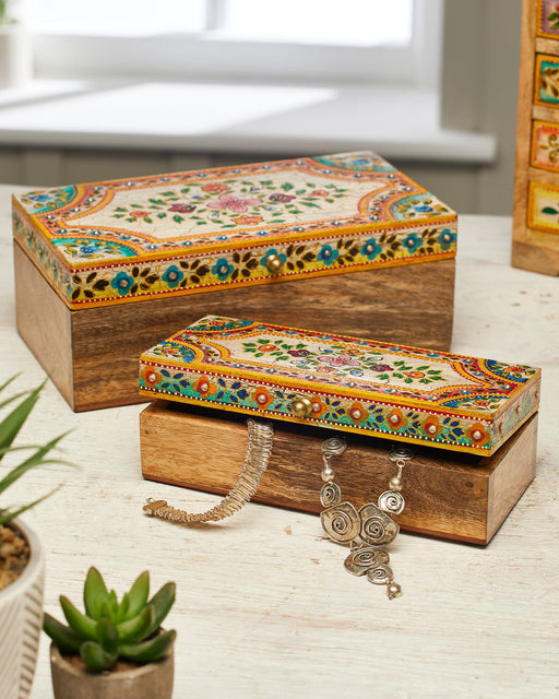 Decorative wooden boxes with floral patterns on a light surface, with a plant in the foreground.