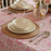 A close-up view of a place setting on a pink floral block-print tablecloth. The setting includes a stacked cream-colored bowl and plate on a round braided jute charger. Silver cutlery and a wine glass are positioned beside the plate, with soft candlelight and a rose centerpiece blurred in the background.