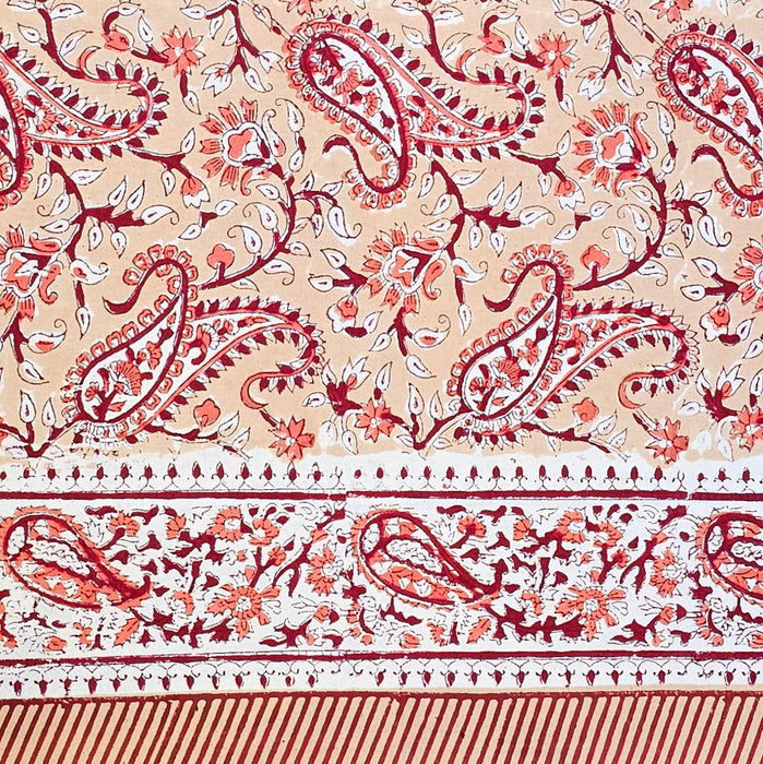 A close-up of a printed cotton tablecloth featuring red and orange paisley and floral motifs on a warm peach background, with a decorative border running horizontally across the lower portion.